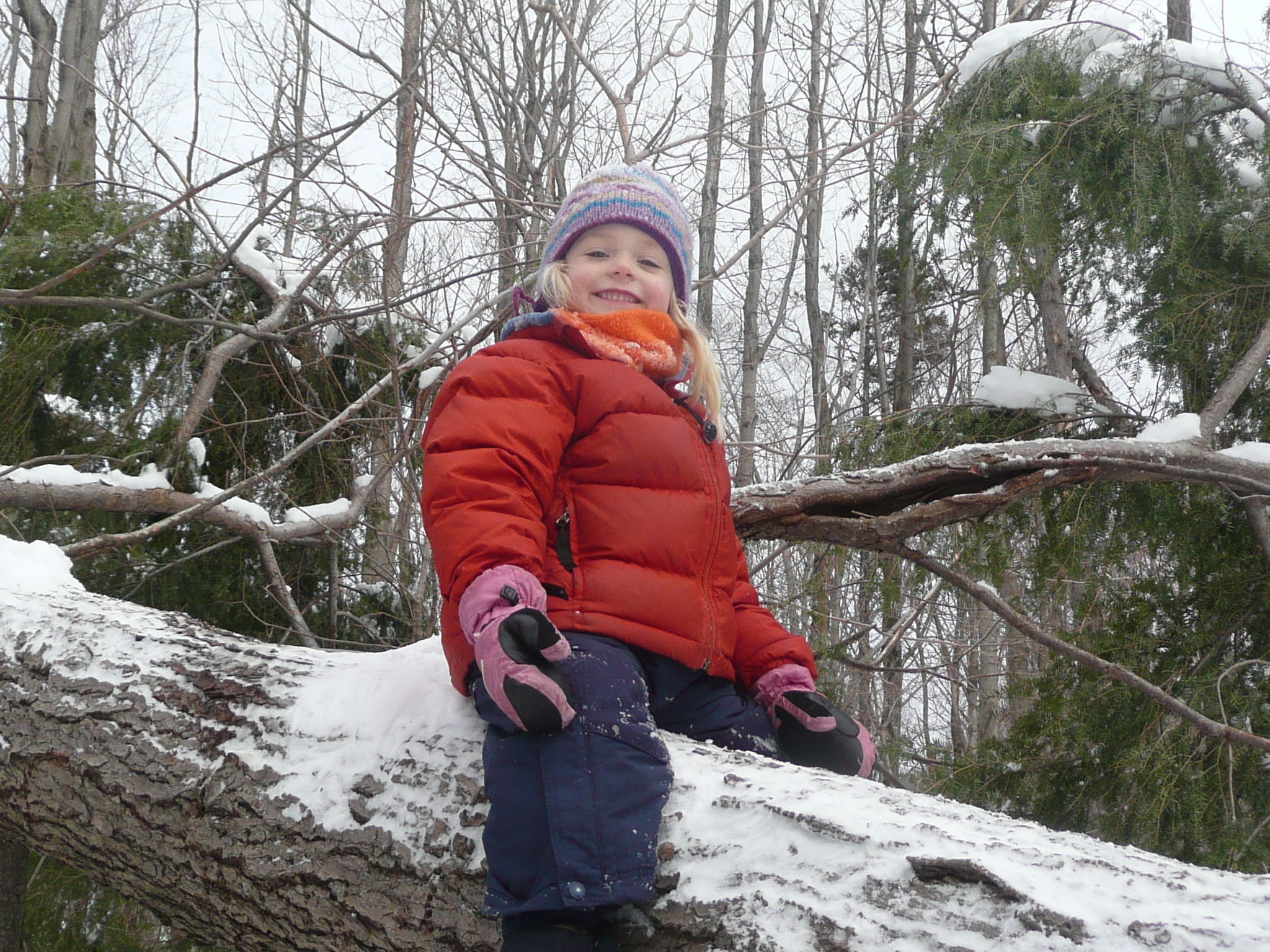 A young child in red puffy coat sit and smiles on a snow covered log