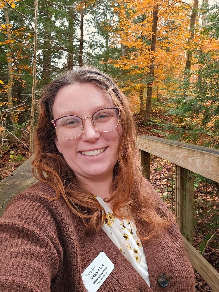 Office Coordinator Meghan Lee-Hall, standing on a wooden bridge with fall foliage in the background.