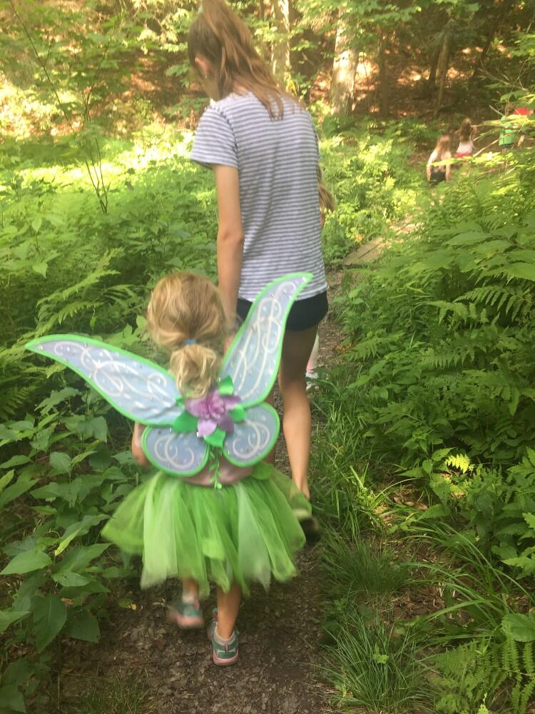 A young camper being led down a fern bordered trail by a camp counselor. The camper is wearing green fairy wings and a green tutu.