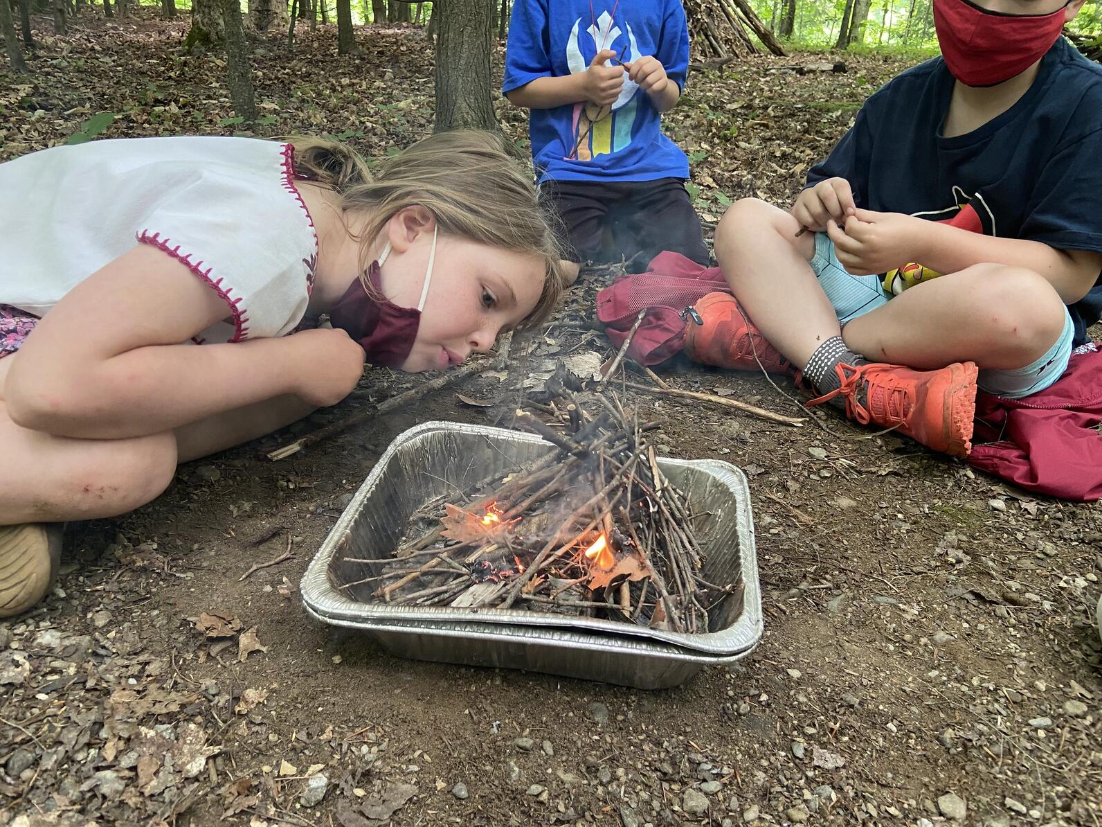 campers work together to keep a small stick fire burning. one camper pulls down their mask to blow on the flames while other look on.