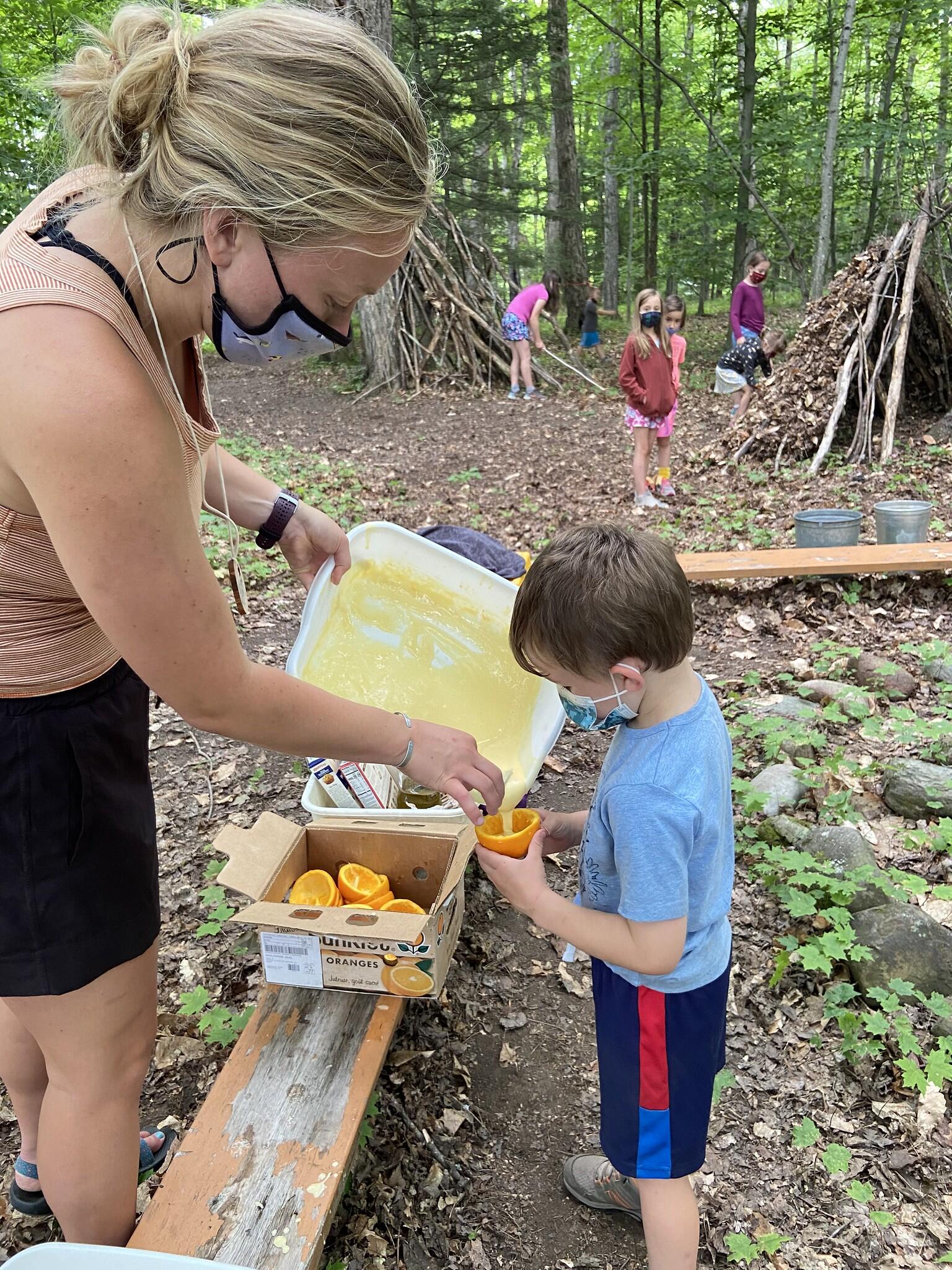 Counselor Mady helps a camper pour muffin mix into a half-orange peel
