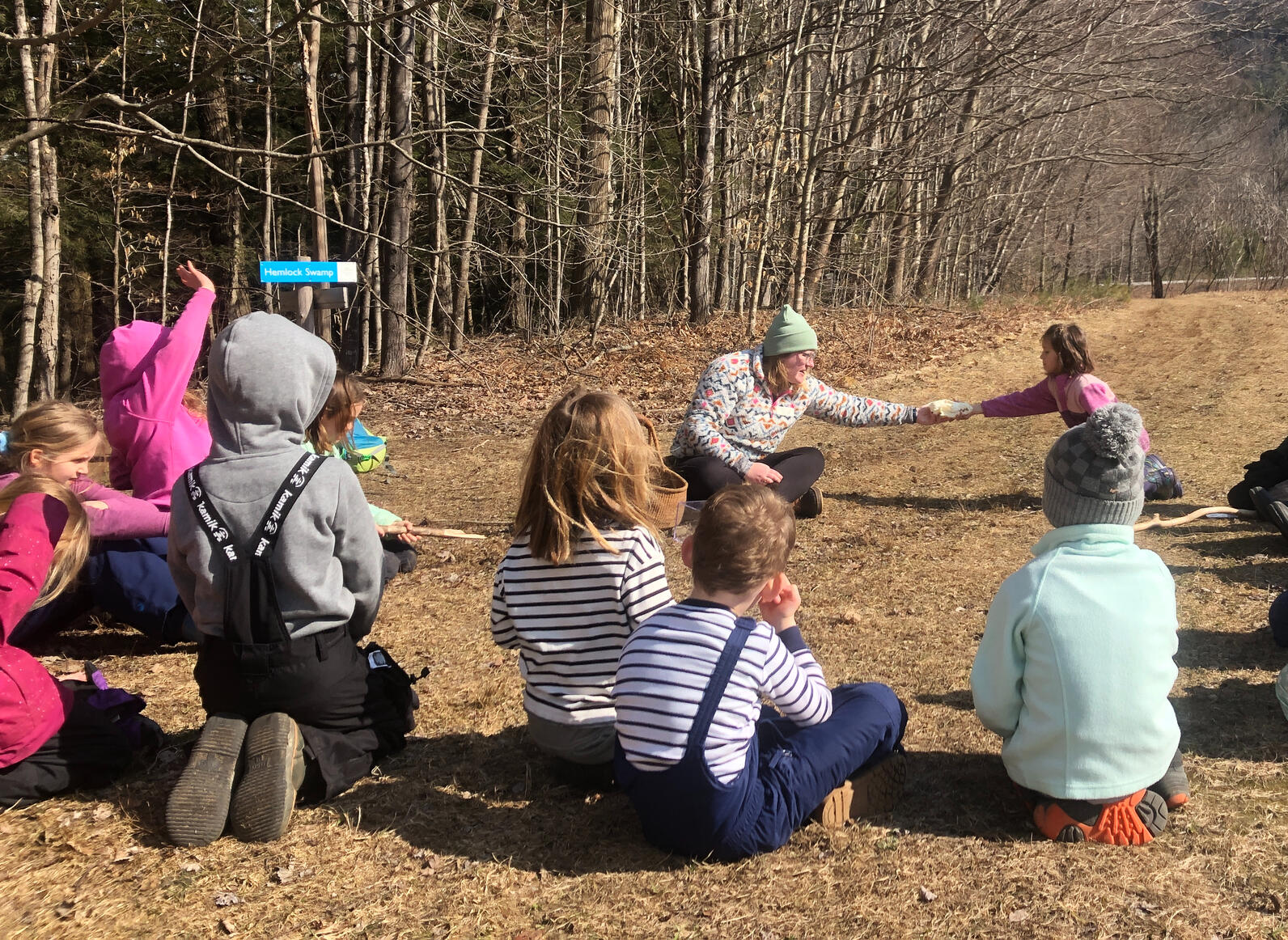 Children sit in a circle in the grass. On the far side of hte circle a teacher passes a skull to the child next to her.