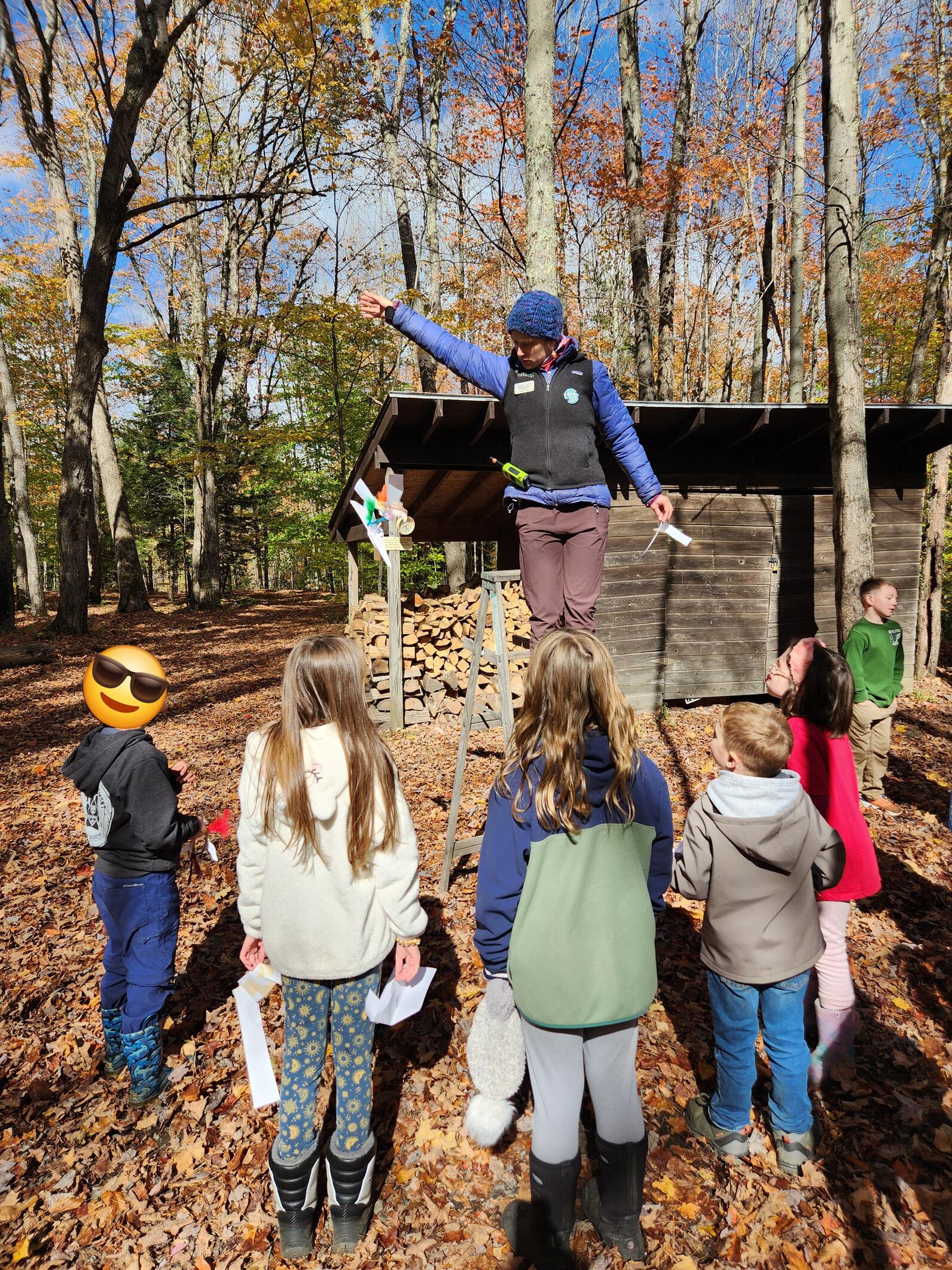 Several kids gather around a teacher on a step ladder watching as she drops an engineered seed made of paper and feathers