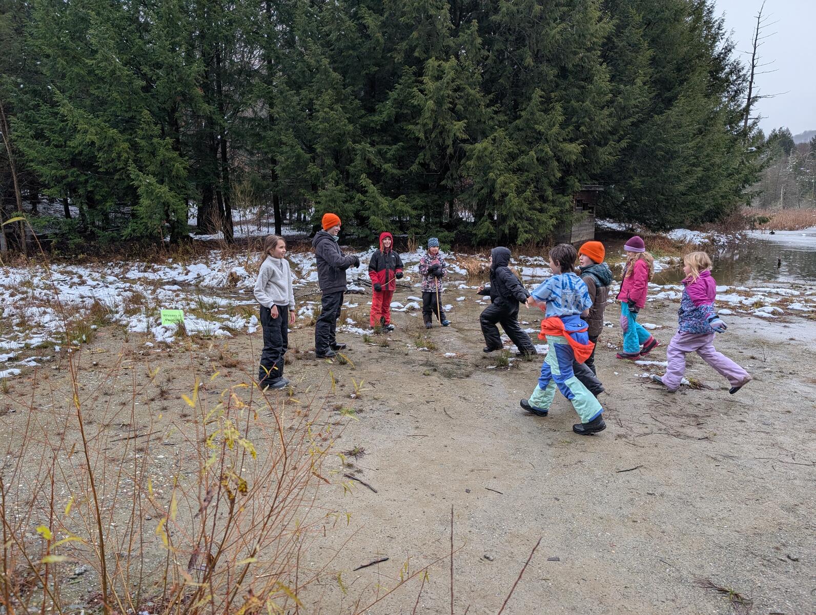 A group of kids stand in a loose clump on a mud flat next to a pond. They are moving around. On the ground is a sign that says "stays in vermont"