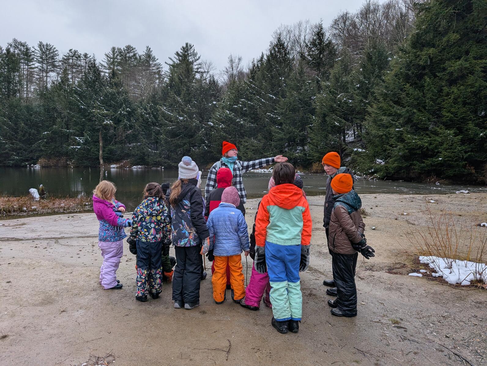 A cluster of students stand by a teacher as she talks to them. They are standing on a mud flat with a pond in the background.