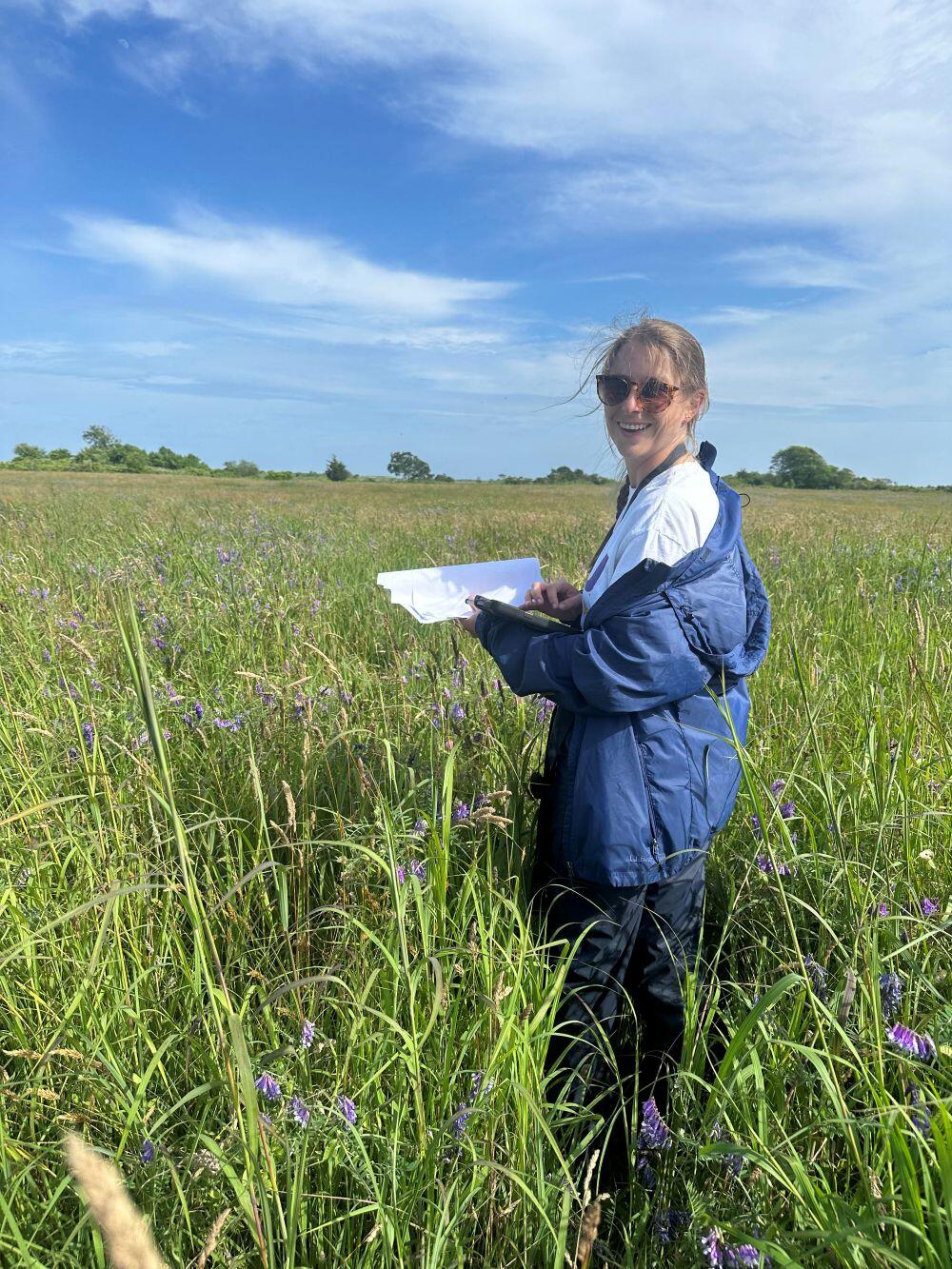 A photo of Macie standing in a tall grassy field holding notes and smiling at the camera. 