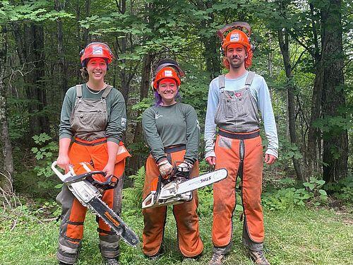 Three youth, wearing hardhats, face shields, and orange safety pants hold chainsaws against a forest backdrop.
