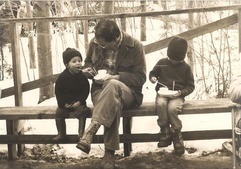 A photo of a parent sitting on a fence with two children, eating sugar on snow.