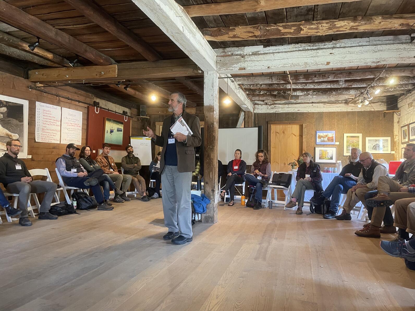 Audubon Vermont's Executive Director David Mears standing in the middle of a large room in a barn. Other Vermont conservation leaders are sitting in chairs circled around the periphery.