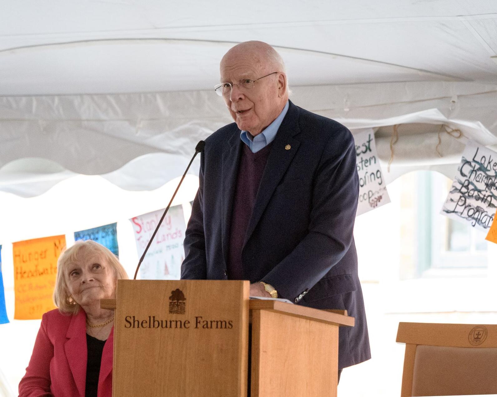 Senator Patrick Leahy, center, stands at a podium that reads “Shelburne Farms,” while Marcelle Pomerleau, his wife, right, looks on. 
