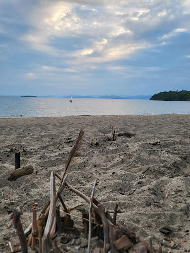 Sticks, pebbles and a feather form a triangle shaped structure on the beach of Lake Champlain. A small sailboat is sailing away on calm water. 