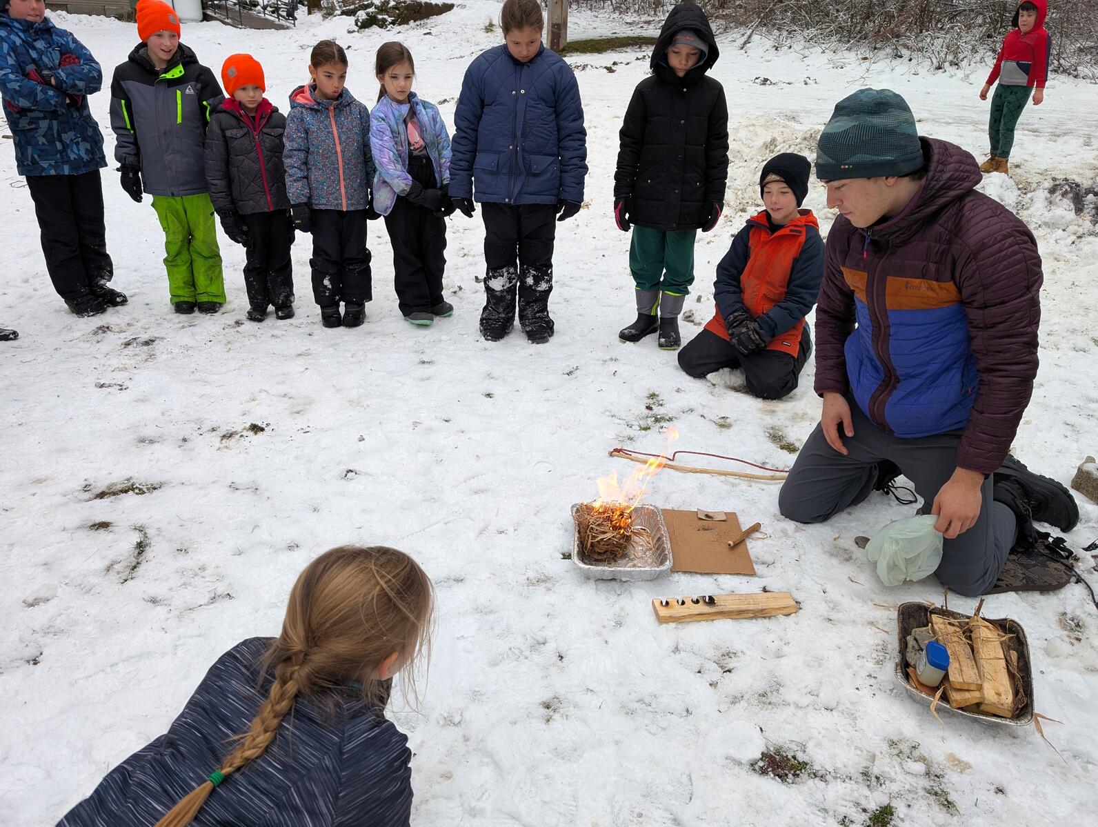 A group of kids stand and watch as a man sits by a freshly made fire in a tray. A bow drill lays to the side of the man A group of kids stand and watch as a man sits by a freshly made fire in a tray. A bow drill lays to the side of the man