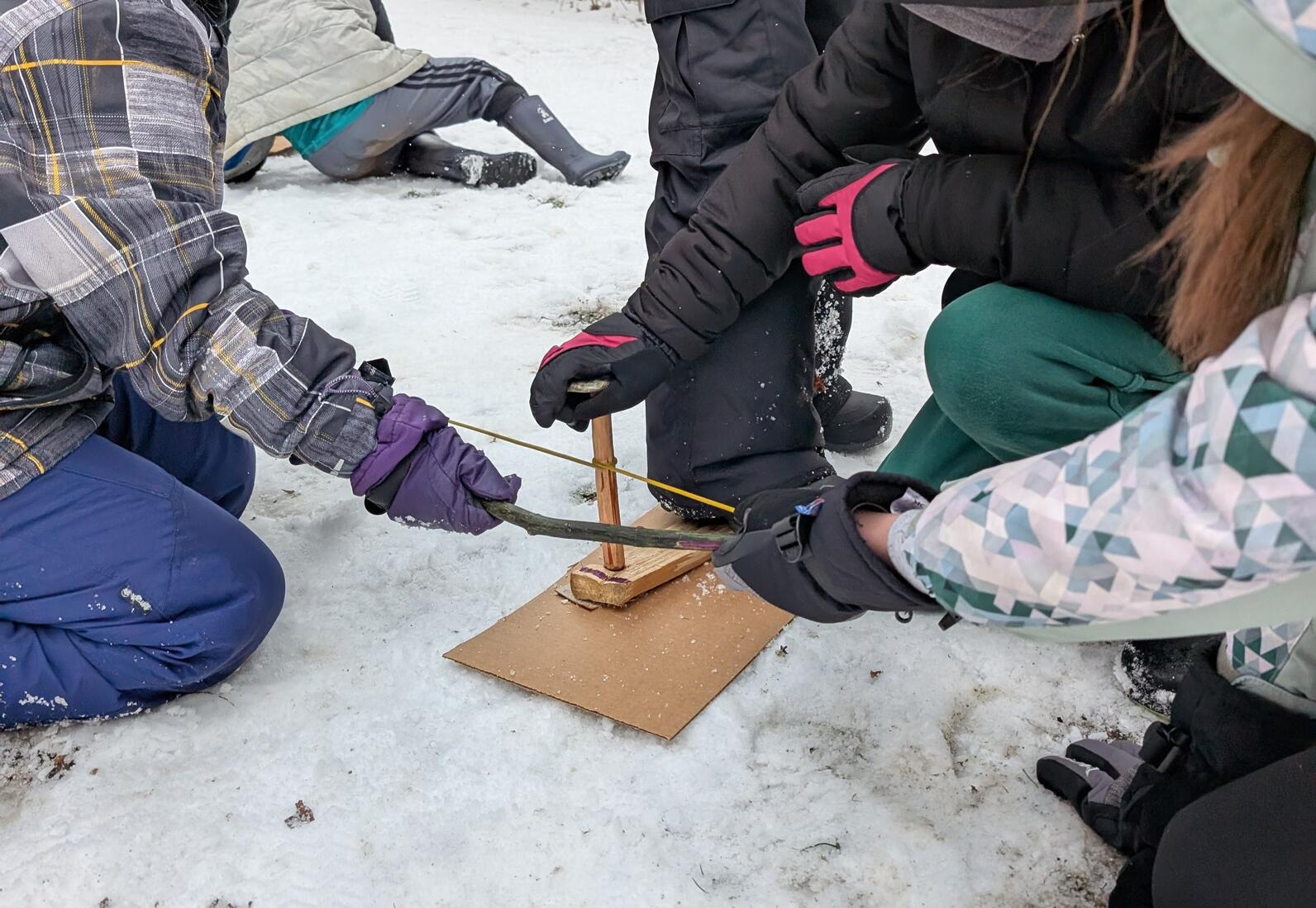 4 kids work together to use a bow drill. One stands on the base board, one holds the stone, and two hold the bow 4 kids work together to use a bow drill. One stands on the base board, one holds the stone, and two hold the bow