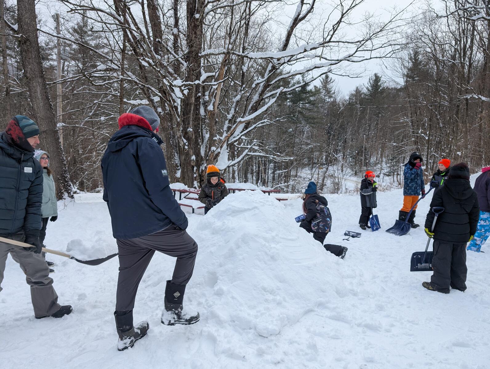A group of children and their teachers are gathered around a pile of snow with shovels in their hands adding snow to the pile A group of children and their teachers are gathered around a pile of snow with shovels in their hands adding snow to the pile