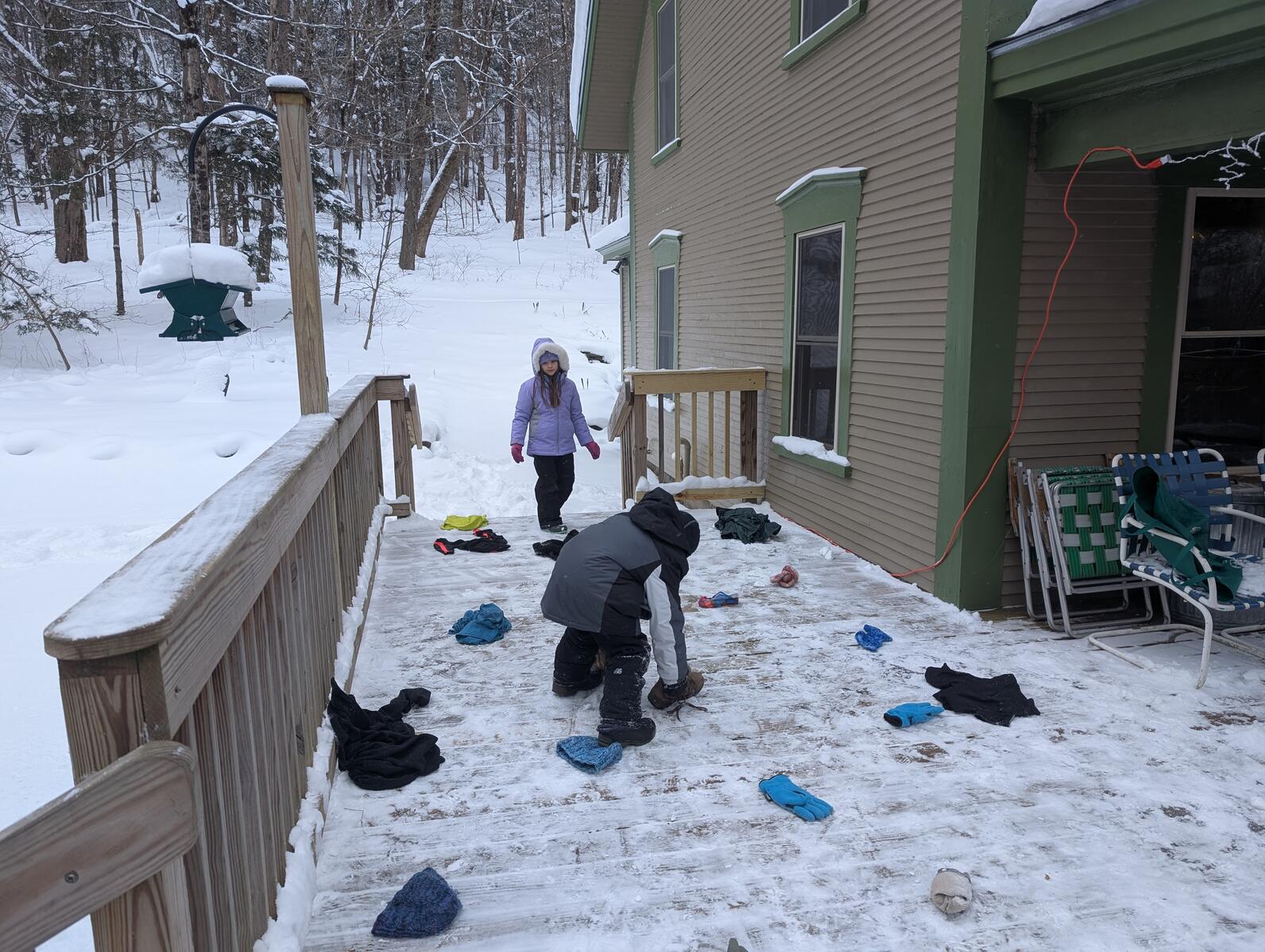 Two kids stand on a porch with pieces of clothing on the ground around them. There is a house on the right that the porch is attached to and a snowy forest behind them. One kid bends down to pick up a boot. Two kids stand on a porch with pieces of clothing on the ground around them. There is a house on the right that the porch is attached to and a snowy forest behind them. One kid bends down to pick up a boot.