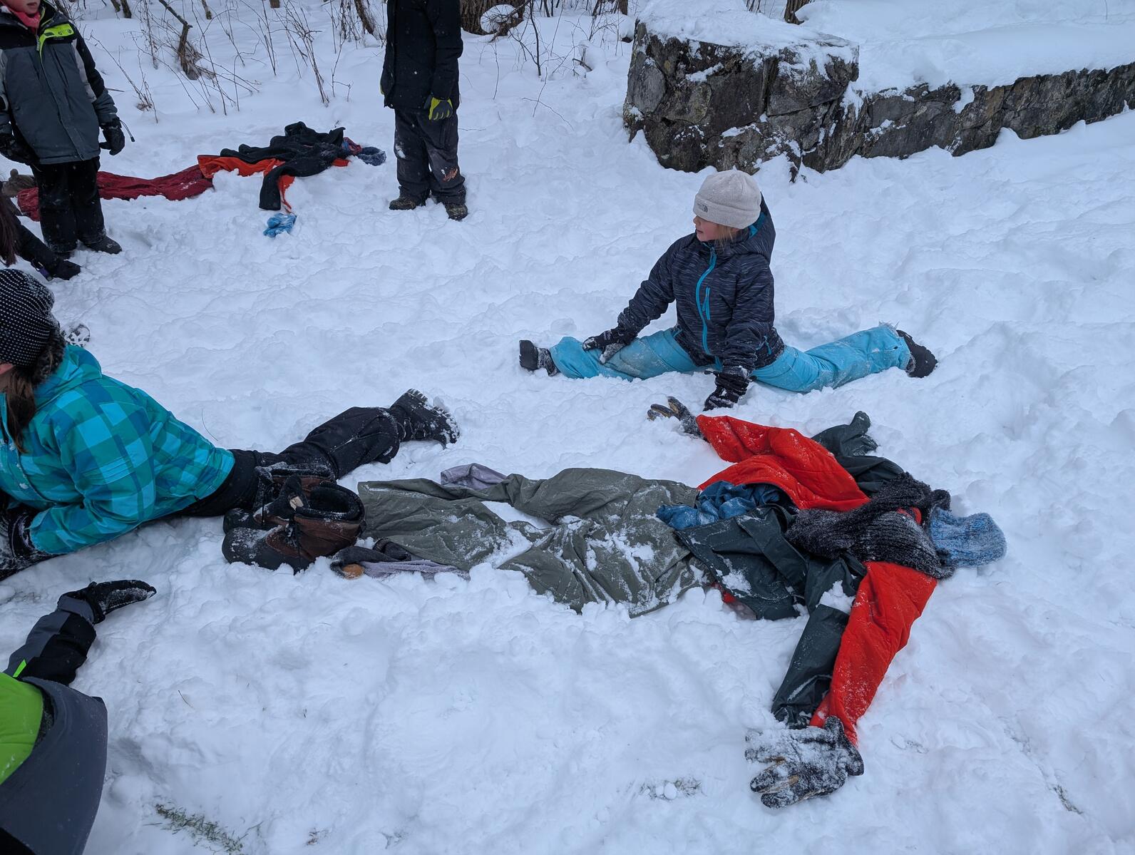 Winter clothes are layed out in the shape of a person in the snow while two girls do splits in the background