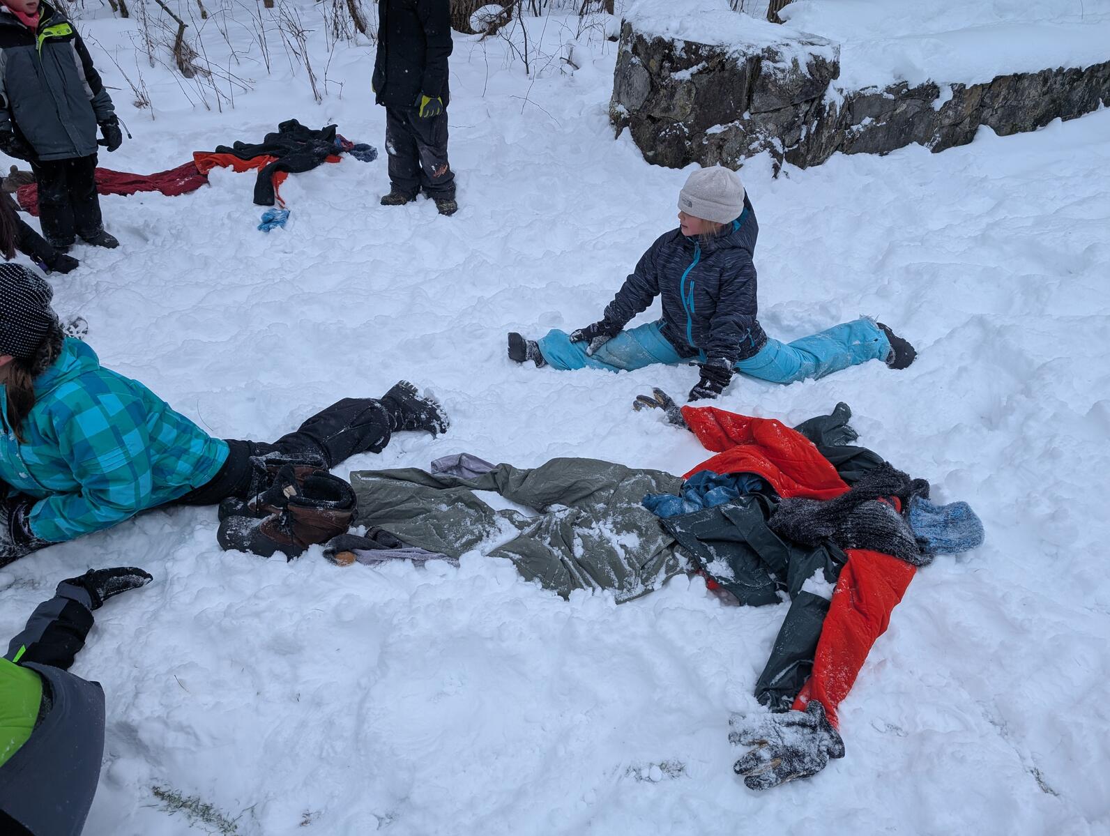 Winter clothing in the outline of a person lay in the snow on the ground in the foreground. Another such pile of clothing is visible in the background. There are two girls doing splits in the snow near the close pile and a child standing near the far pile Winter clothing in the outline of a person lay in the snow on the ground in the foreground. Another such pile of clothing is visible in the background. There are two girls doing splits in the snow near the close pile and a child standing near the far pile