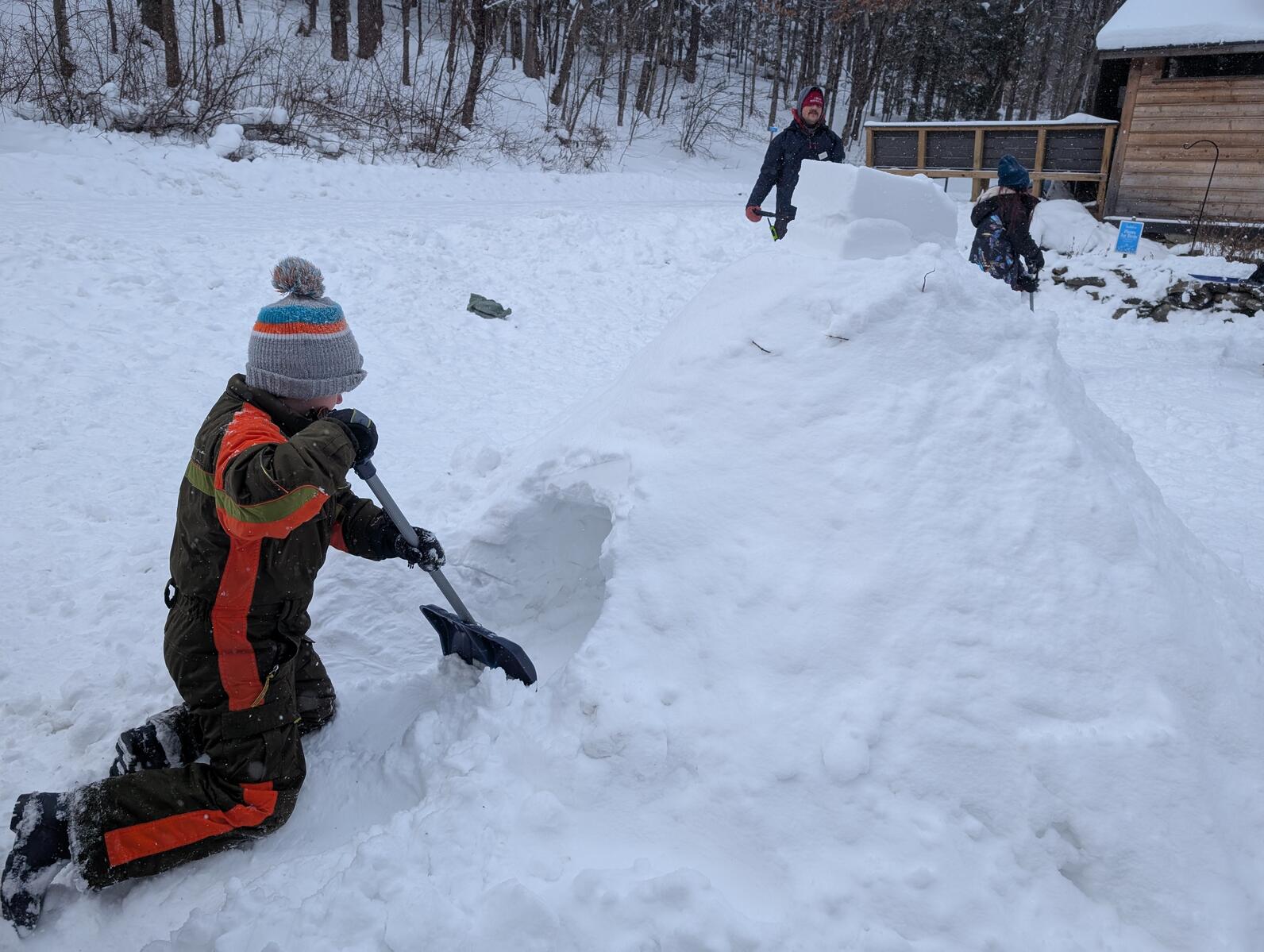 A child in a orange and brown snowsuit kneels in the snow in front of a snow pile. The pile has a hole going through it. The boy holds a shovel scooping show out of the hole A child in a orange and brown snowsuit kneels in the snow in front of a snow pile. The pile has a hole going through it. The boy holds a shovel scooping show out of the hole