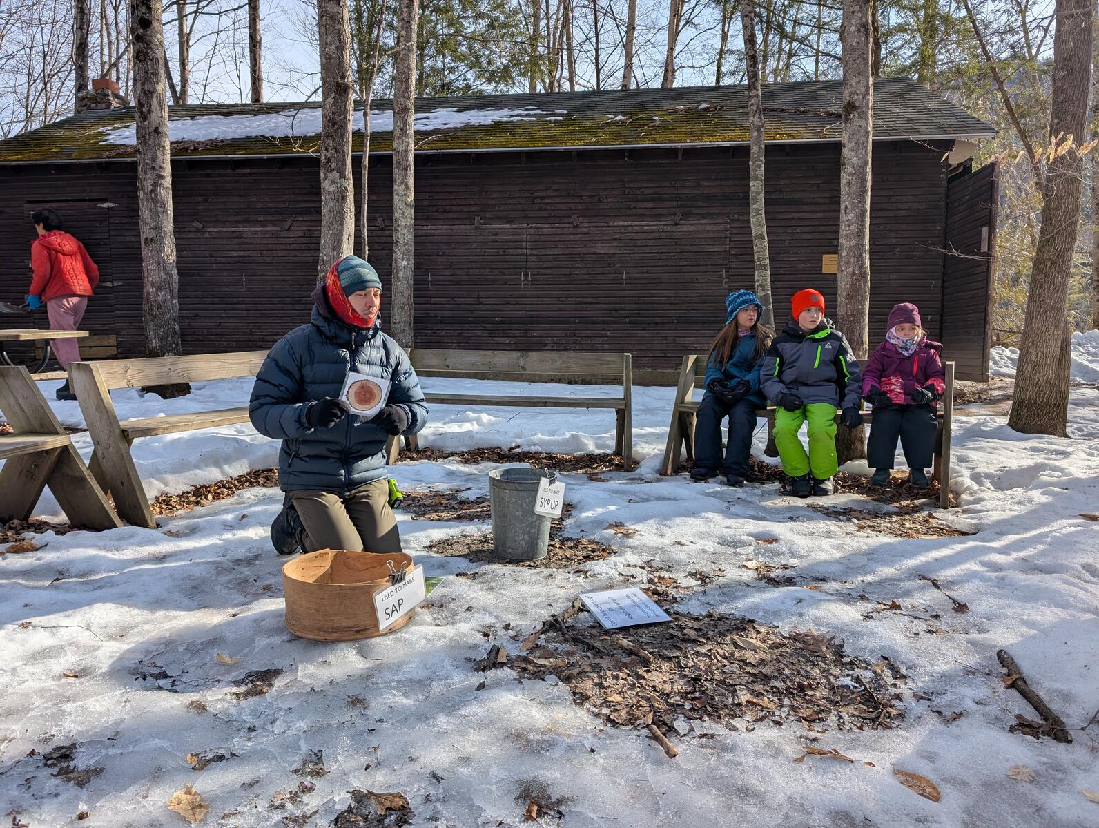 a teacher kneels in the center of a circle of benches holding up an image of the cross cut of a tree trunk. Three kids sit on a bench next to him of a teacher kneels in the center of a circle of benches holding up an image of the cross cut of a tree trunk. Three kids sit on a bench next to him of