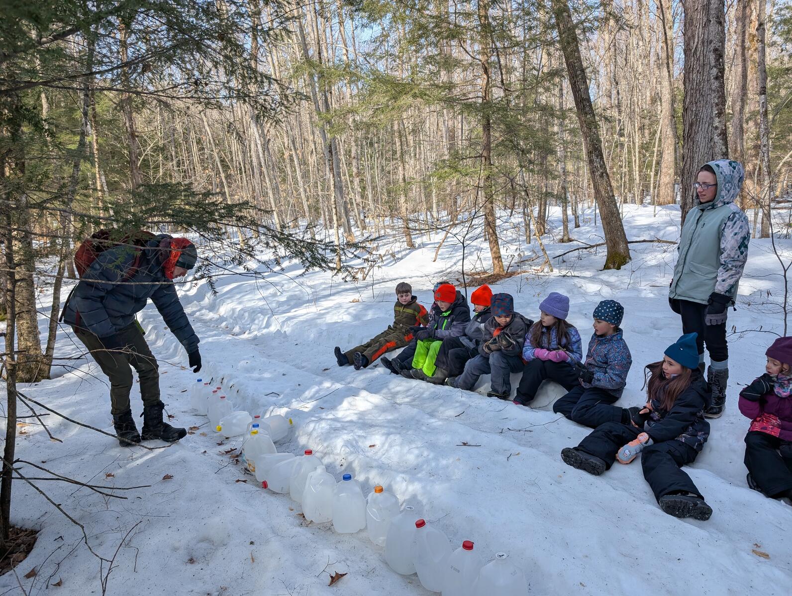 About a dozen students sit in the snow along the edge of a trail while a teacher counts a line of empty milk jugs on the trail About a dozen students sit in the snow along the edge of a trail while a teacher counts a line of empty milk jugs on the trail