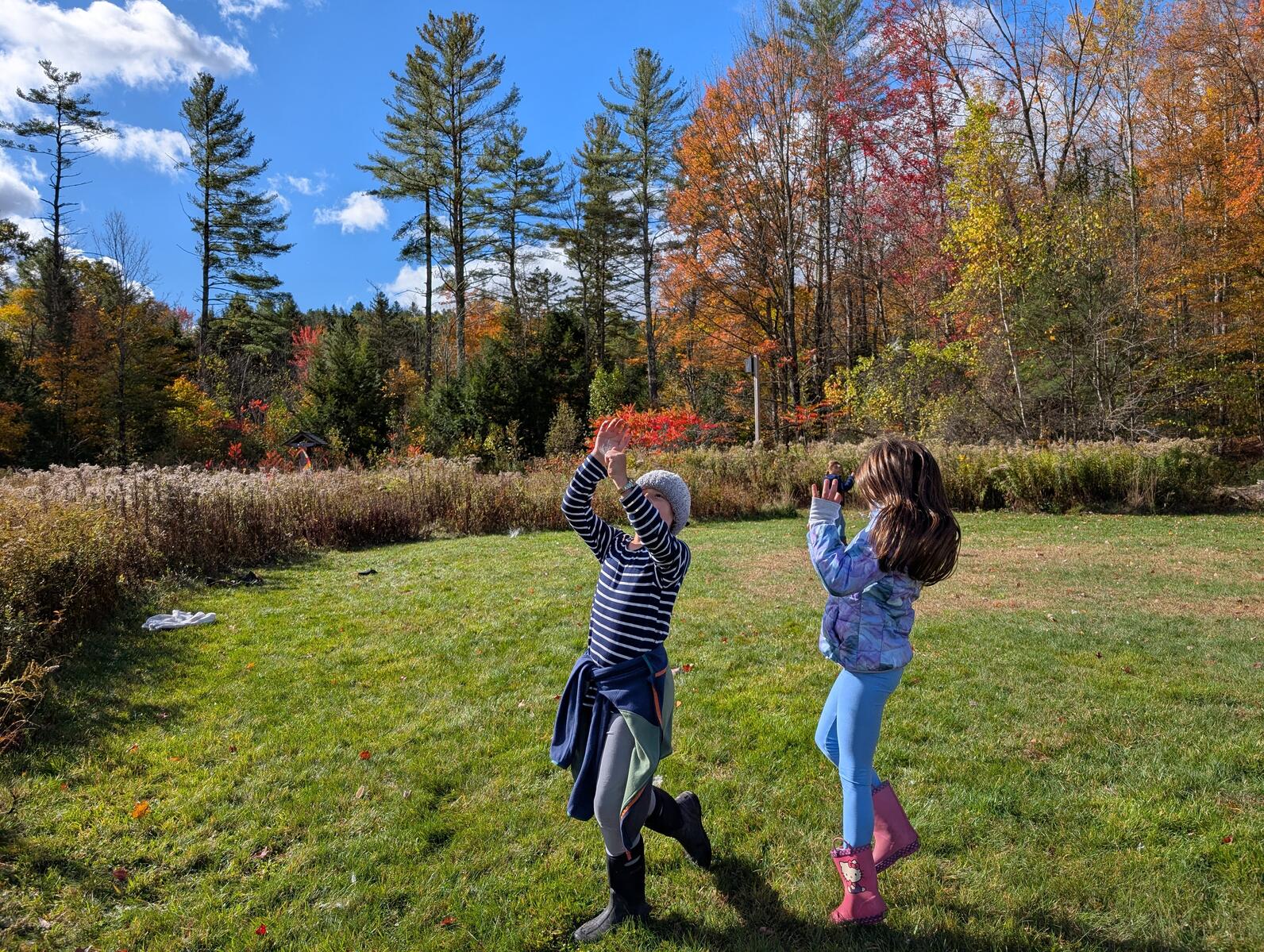 Two girls skip and spin in a field of short grass surrounded by tall grass and colorful trees with their hands in the air catching milkweed seeds