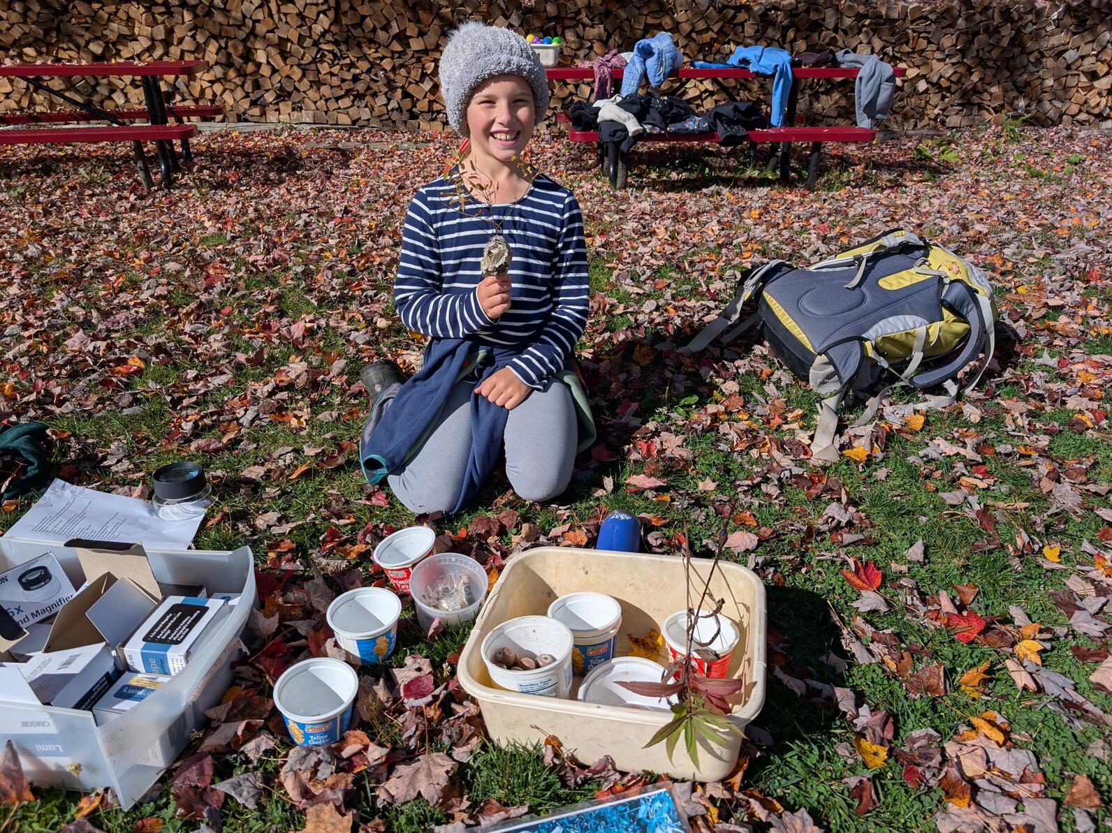Girl sitting in the grass behind small yogurt cups filled with seeds and other nature findings holding a small empty wasp nest