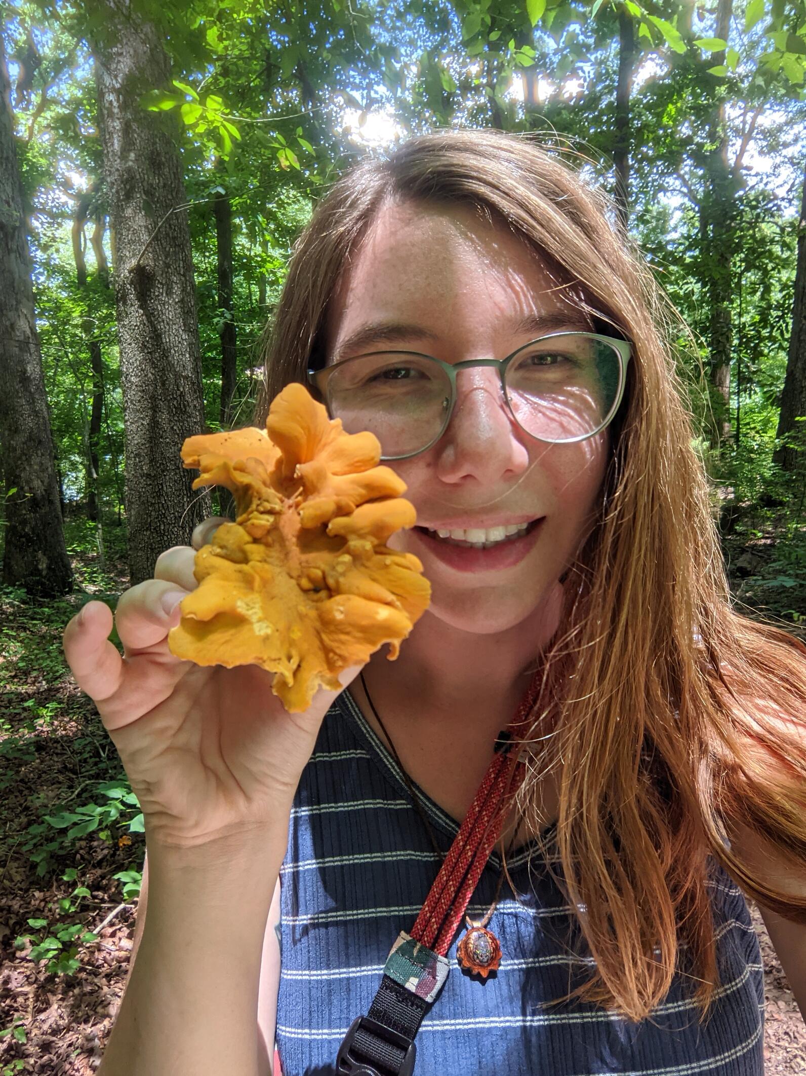 Samantha holds a golden orange colored mushroom close to the camera while standing in the woods on a sunny day wearing a blue stripped tank top.