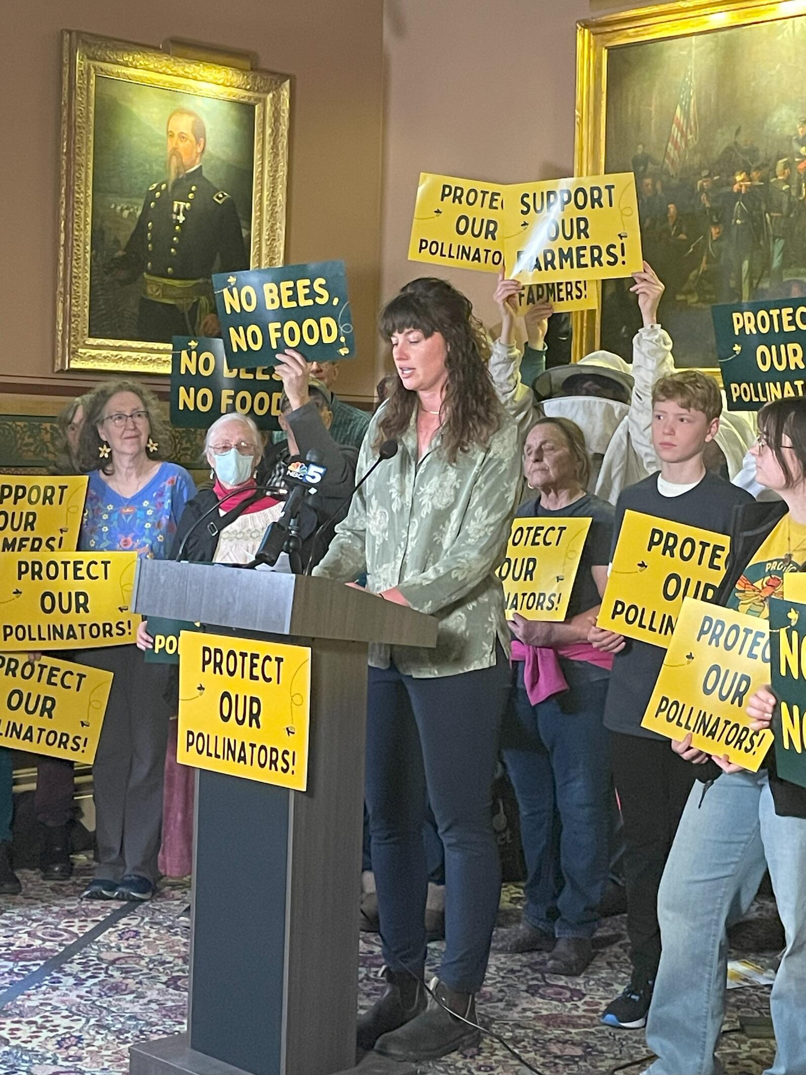 Woman standing at a podium surrounded by people with protect our pollinators signs