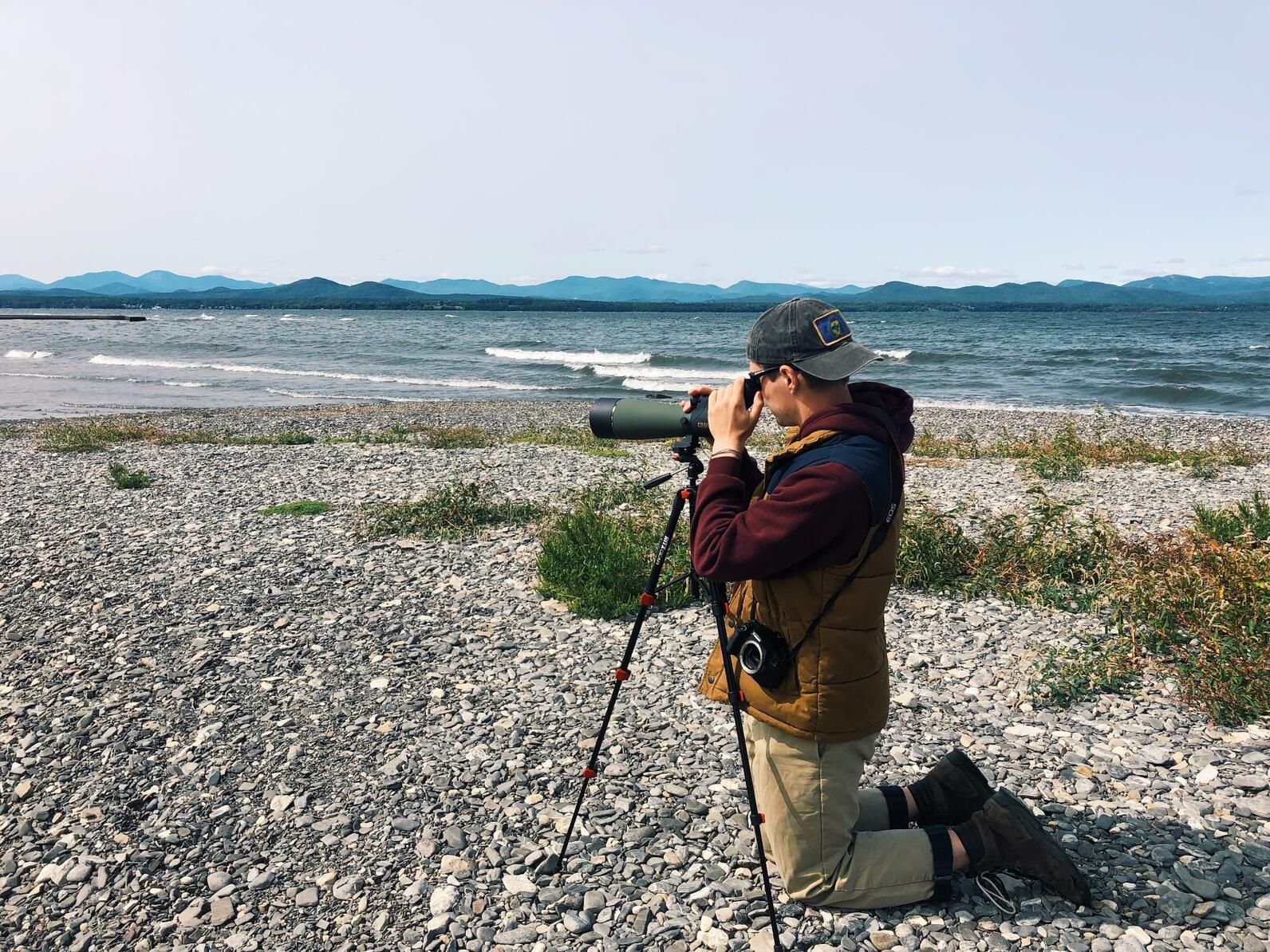 Conservation Intern Tyler Philbrook kneels on a rocky shore to peer into a spotting scope, looking for birds.