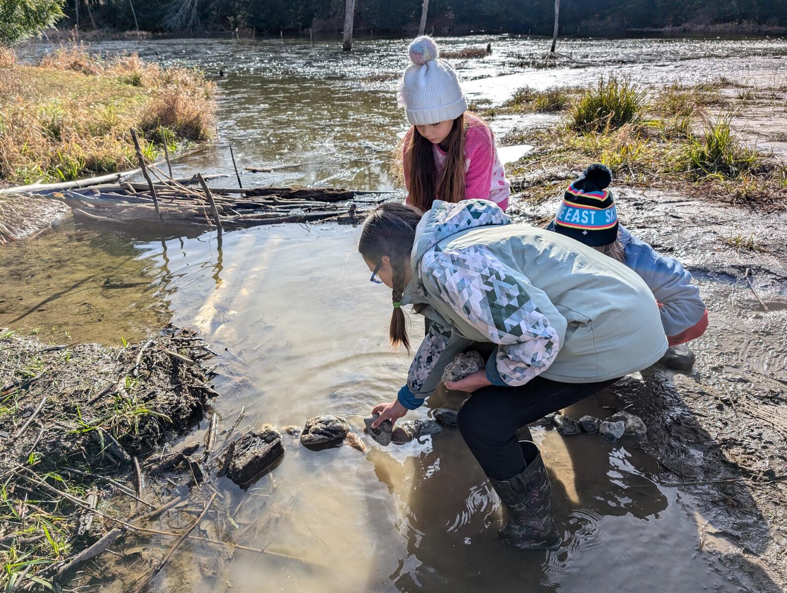 Three girls work together to build a dam out of rocks and mud in a small stream with a pond in the background