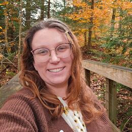 Office Coordinator Meghan Lee-Hall, standing on a wooden bridge with fall foliage in the background.