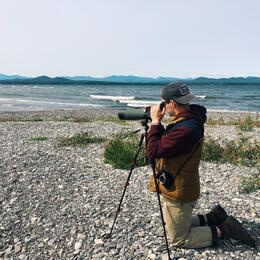 Conservation Intern Tyler Philbrook kneels on a rocky shore to peer into a spotting scope, looking for birds.