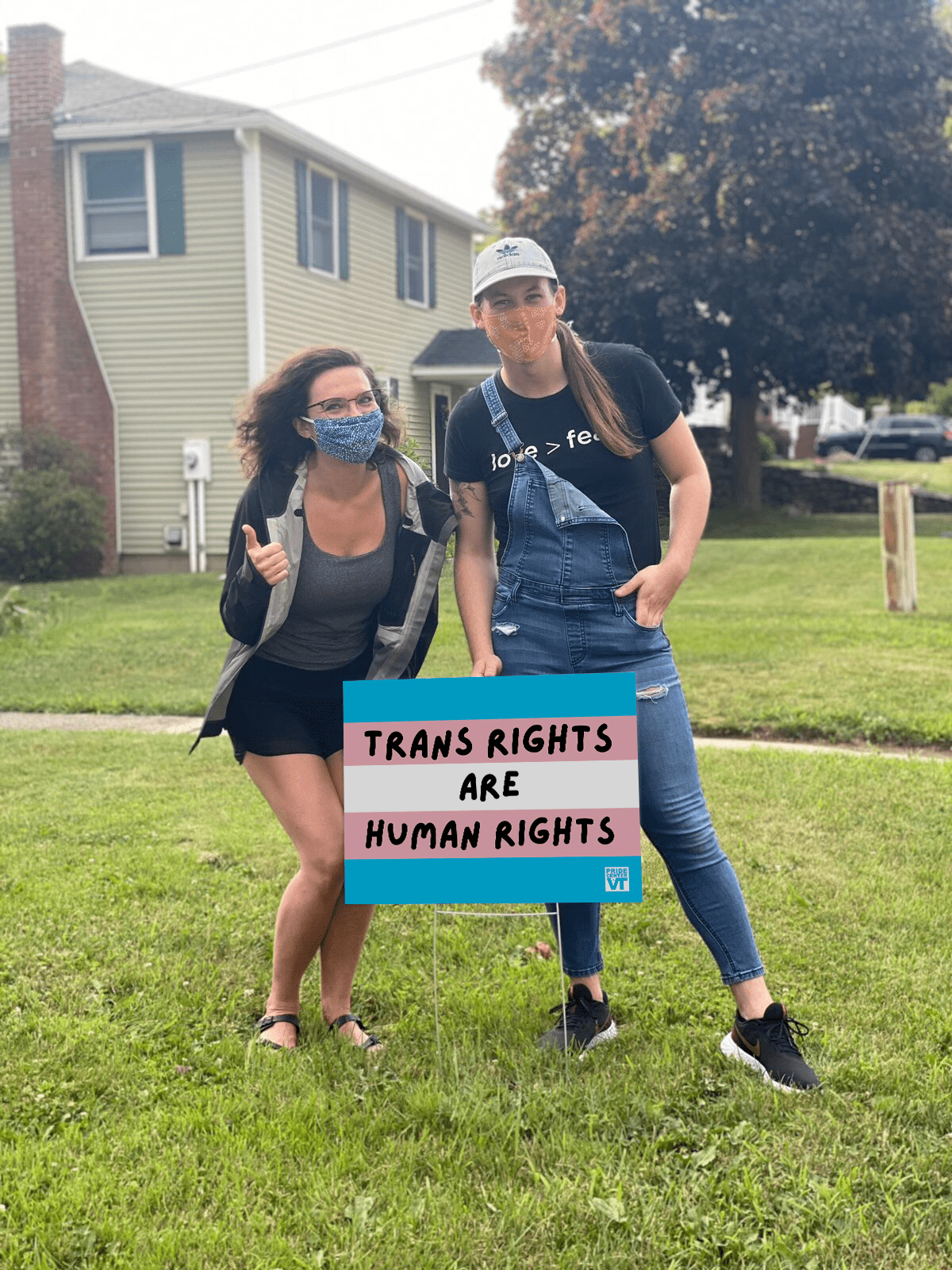 Two people stand with a yard sign that reads "Trans Rights are Human Rights"