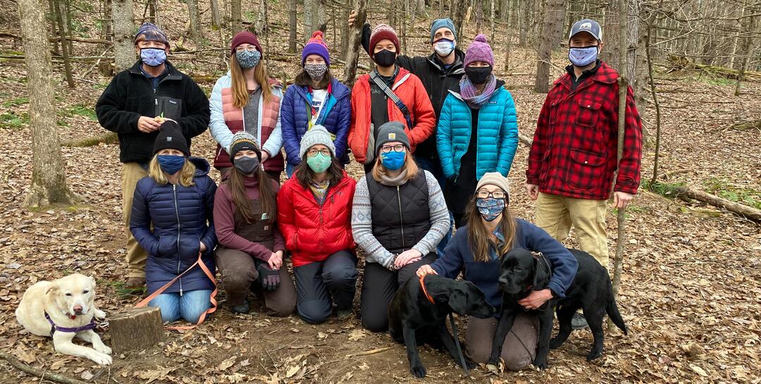 Audubon Vermont Staff photo 2021 - Twelve people, bundled up for winter, and wearing COVID masks pose for a group photo in the winter woods in Vermont. Three dogs join the photo.  