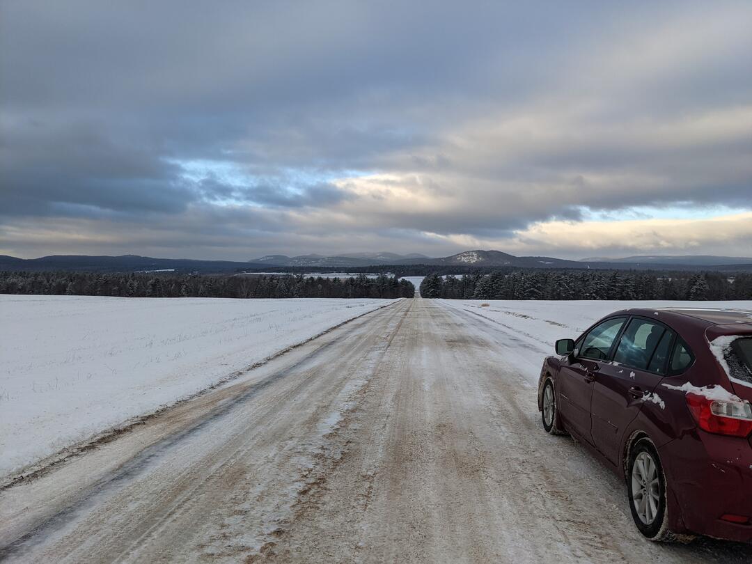 A car stops along the side of a snow-covered road. Vermont's Green Mountains are visible in the distance. Snow-covered farm fields extend in both directions around the road.